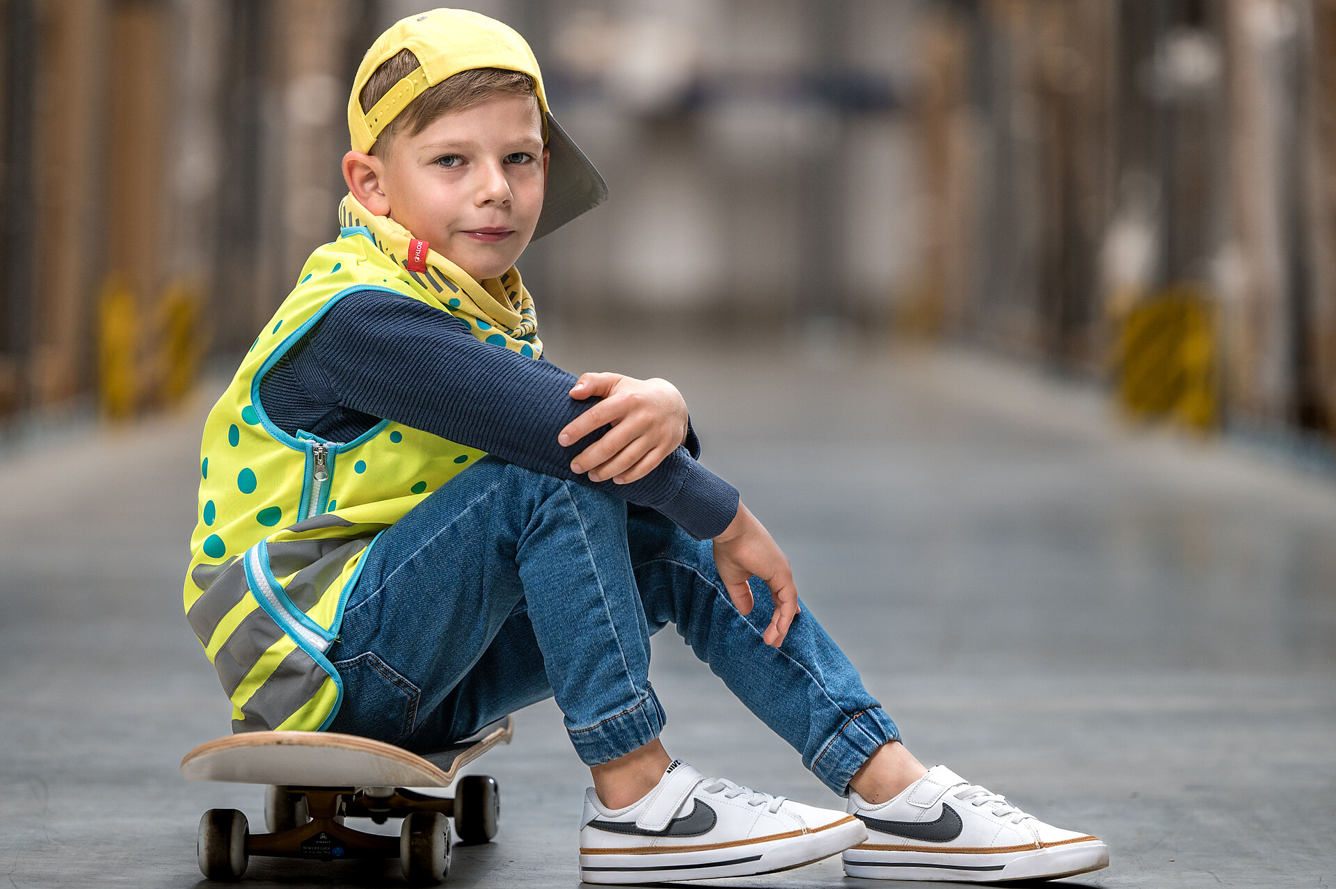 Junge mit gelber Warnweste und Basecap sitzt auf einem Skateboard